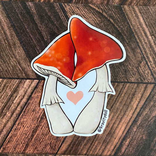 A die cut sticker of two red mushrooms leaning toward one another. The mushrooms frame a pink heart floating between them.