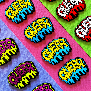 The red and white Queer enamel pin is showcased with other color varieties. Goopy white letters spell out "QUEER" with additional red background goop dripping downward.