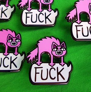 Closeup of many Foul Meowthed Enamel pins. An angry, bristled pink cat stands atop a giant speech bubble, saying "FUCK" in all caps.