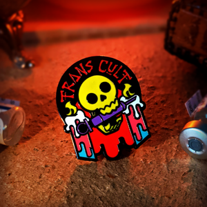 The trans cult enamel pin is black cast and depicts a skull munching on a purple syringe, fixed between two burning candles. TRANS CULT is written in red across the top of the pin.