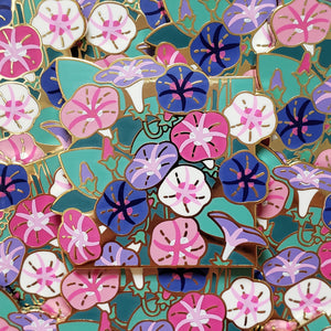 Closeup of the gold cast pin which features a bundle of morning glory flowers. The flowers are the colors of the genderfluid flag (white, pink, blue, purple, and black)).