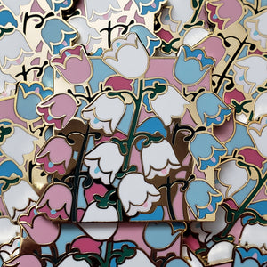 Closeup of the gold cast pin which features a bundle of canterbury bells flowers. The flowers are the colors of the trans flag (pastel pink, white, and pastel blue).