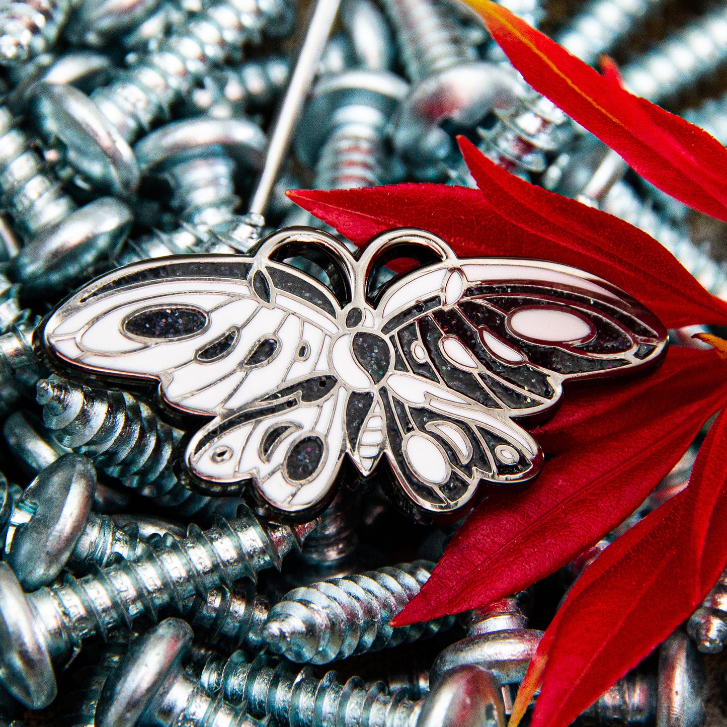 White and sparkling black Iron Widow butterfly enamel pin on pile of screws with red flower.