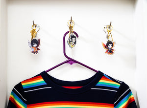 All three Iron Widow charms, attached to house keys and hanging from brass wall hooks with a hanging rainbow shirt on a purple hanger beneath them.