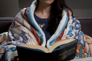 Photo of a person from the chin to the waist, wrapped in a thick blnket and holding a Tamora Pierce novel.