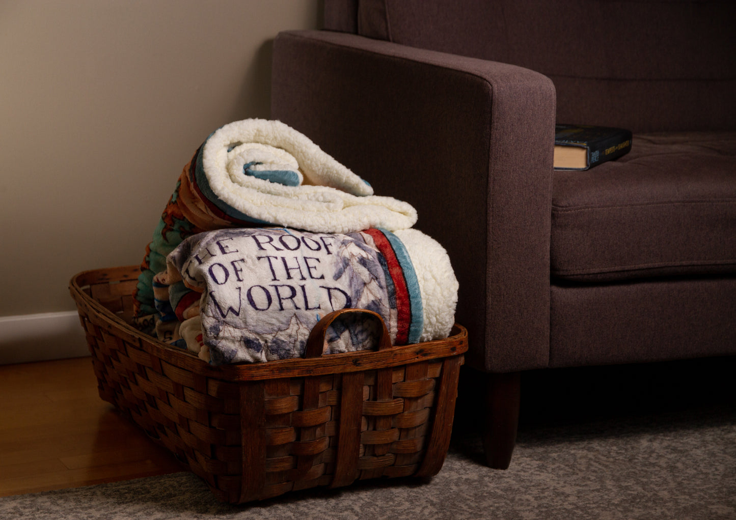 A folded Tortall blanket inside a wicker basket next to a couch. "THE ROOF OF THE WORLD" portion of the illustration is visible on the blanket.