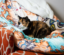 Load image into Gallery viewer, Close-up of the Tortall blanket draped over a chair. The illustration is obscurred by a very good calico cat taking a nap.