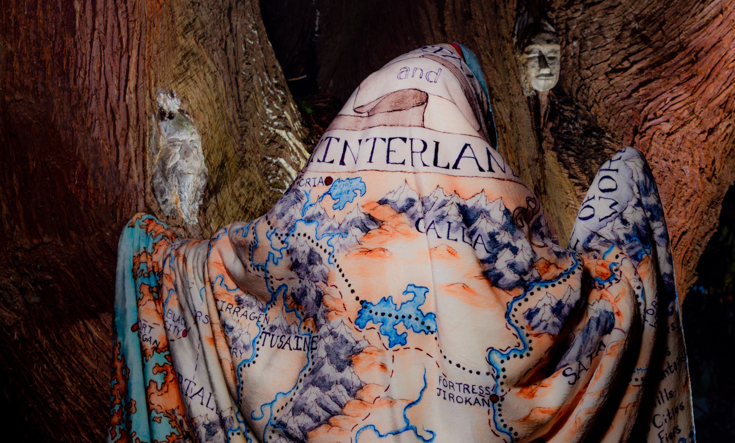 From behind, a person wears the Tortall blanket like a spooky ghost wandering in an equally spooky forest. Much of the illusstration is visible.