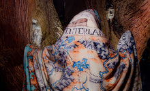 Load image into Gallery viewer, From behind, a person wears the Tortall blanket like a spooky ghost wandering in an equally spooky forest. Much of the illusstration is visible.