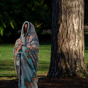 A person stands outside next to a large tree while wrapped head to toe in the Tortall blanket.