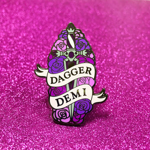The goldcast pin features a decorative dagger in a bed of roses in the purple and pink colors of the demi flag. A ribbon trails across the front and reads "Dagger Demi."