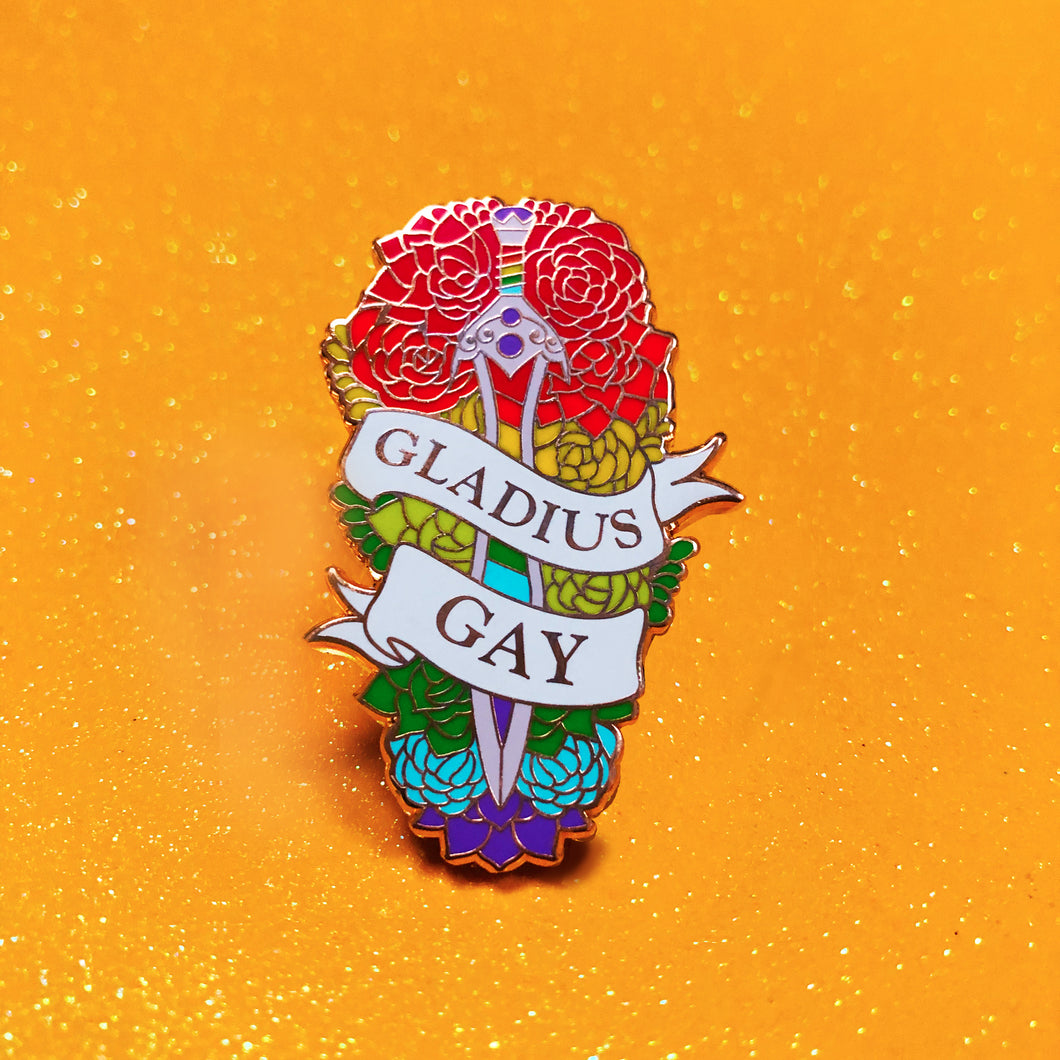 The goldcast pin features a gladius sword in a bed of flowers in rainbow pride colors. A ribbon trails across the front and reads 