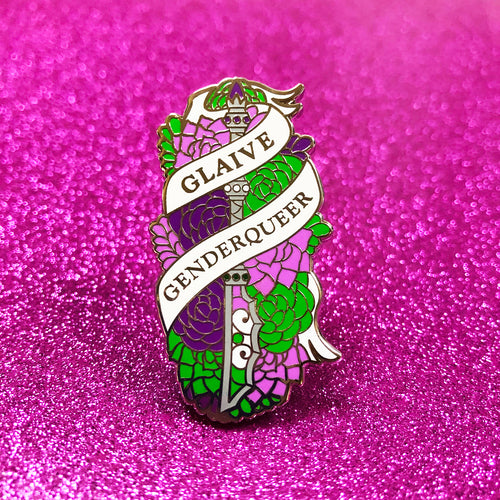 The goldcast pin features a glaive in a bed of roses in the green and purple colors of the genderqueer flag. A ribbon trails across the front and reads 