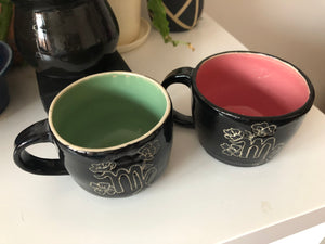 Black Virgo mugs with mint green and pink inside washes sit on a white coffee table. Mugs have Virgo symbol on them surrounded by sprouting flowers.
