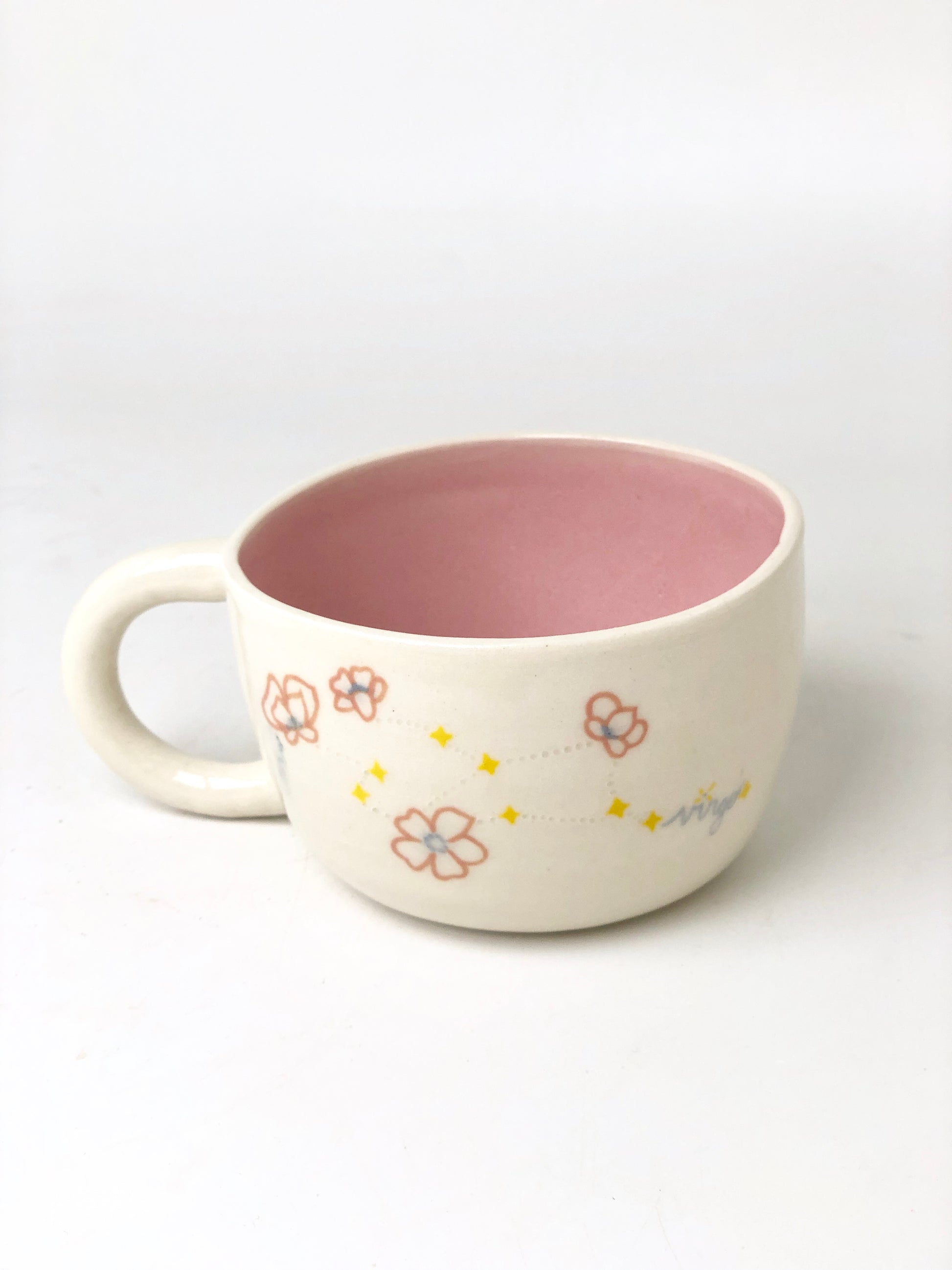White mug with light pink inside and light blue, pink, and yellow star and flower details. Cursive small light blue text on the mug reads "Virgo."
