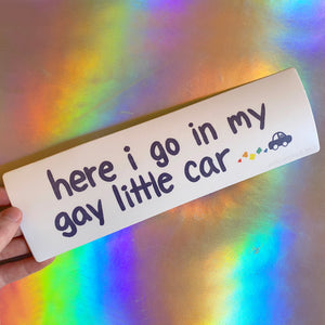 A white bumper sticker with dark purple text reads "here i go in my gay little car" in cartoony font with a small car pooting out rainbow exhaust.