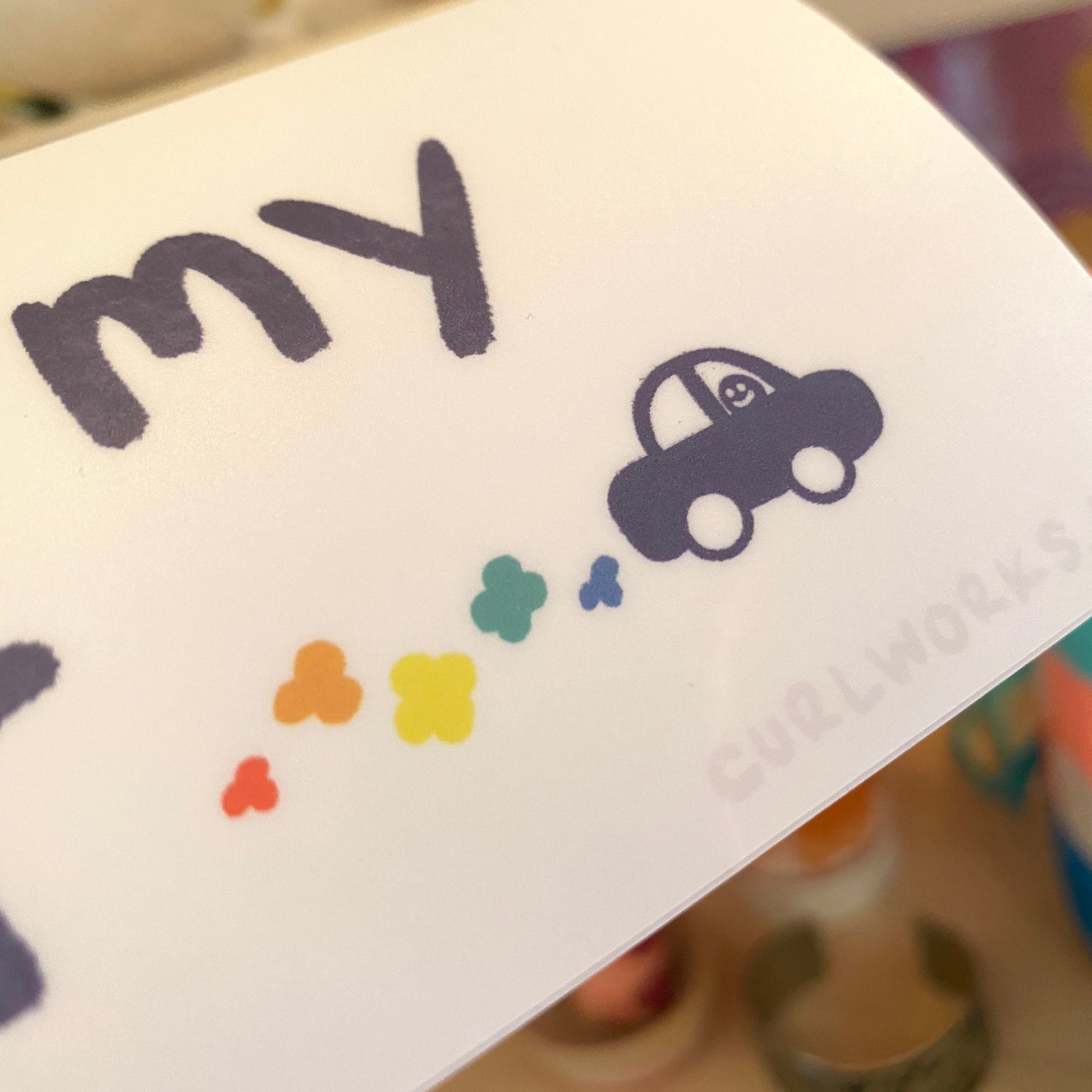 A closeup of the tiny car which shows a small figure with a smiley face driving along with rainbow exhaust trailing out of the car.