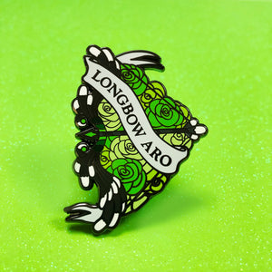 The goldcast pin features a feathery black and white bow in a bed of roses in the green colors of the aromantic flag. A ribbon trails across the front and reads "Longbow Aro"