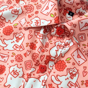 A coral pink button up shirt with heart-shaped red buttons. The shirt features a white and red printed pattern of stylized cute cats with bows holding letters in their mouths, basking among letters, and heart stamps.