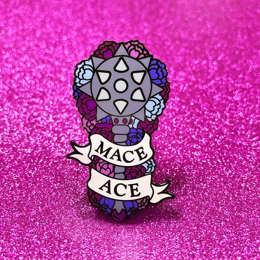 The goldcast pin features a mace weapon in a bed of purple, dark magenta, light blue and pink flowers, representing the colors of the asexual flag. A ribbon trails across the front and reads "Mace Ace."