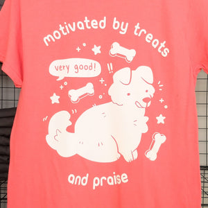 Coral pink t-shirt with a white printed cartoon dog surrounded by bone treats, stars, and sparkles with a speech bubble indicating and unseen speaker saying "very good!" White text surrounding the dog says "motivated by treats and praise."