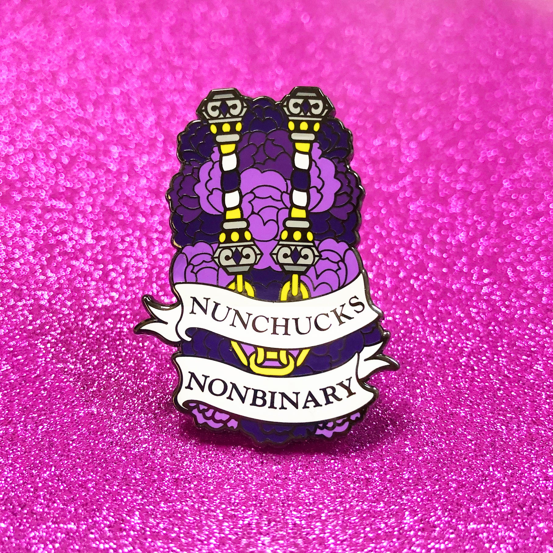The goldcast pin features a pair of purple, white, and bright yellow nunchucks in a bed of purple flowers, representing the nonbinary flag colors. A ribbon trails across the front and reads "Nunchucks Nonbinary."