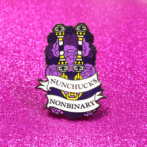 The goldcast pin features a pair of purple, white, and bright yellow nunchucks in a bed of purple flowers, representing the nonbinary flag colors. A ribbon trails across the front and reads "Nunchucks Nonbinary."