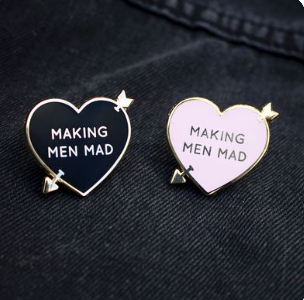 Pink and black variations of gold arrow-pierced, heart-shaped enamel pins with "Making Men Mad" written in the hearts.