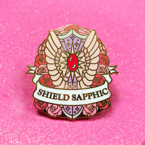 The goldcast pin features a decorative feathery, sparkly shield in a bed of roses in the orange, cream, and pink colors of the sapphic flag. A ribbon trails across the front and reads 