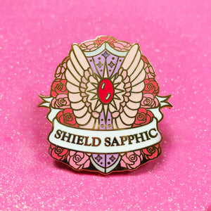 The goldcast pin features a decorative feathery, sparkly shield in a bed of roses in the orange, cream, and pink colors of the sapphic flag. A ribbon trails across the front and reads "Shield Sapphic."