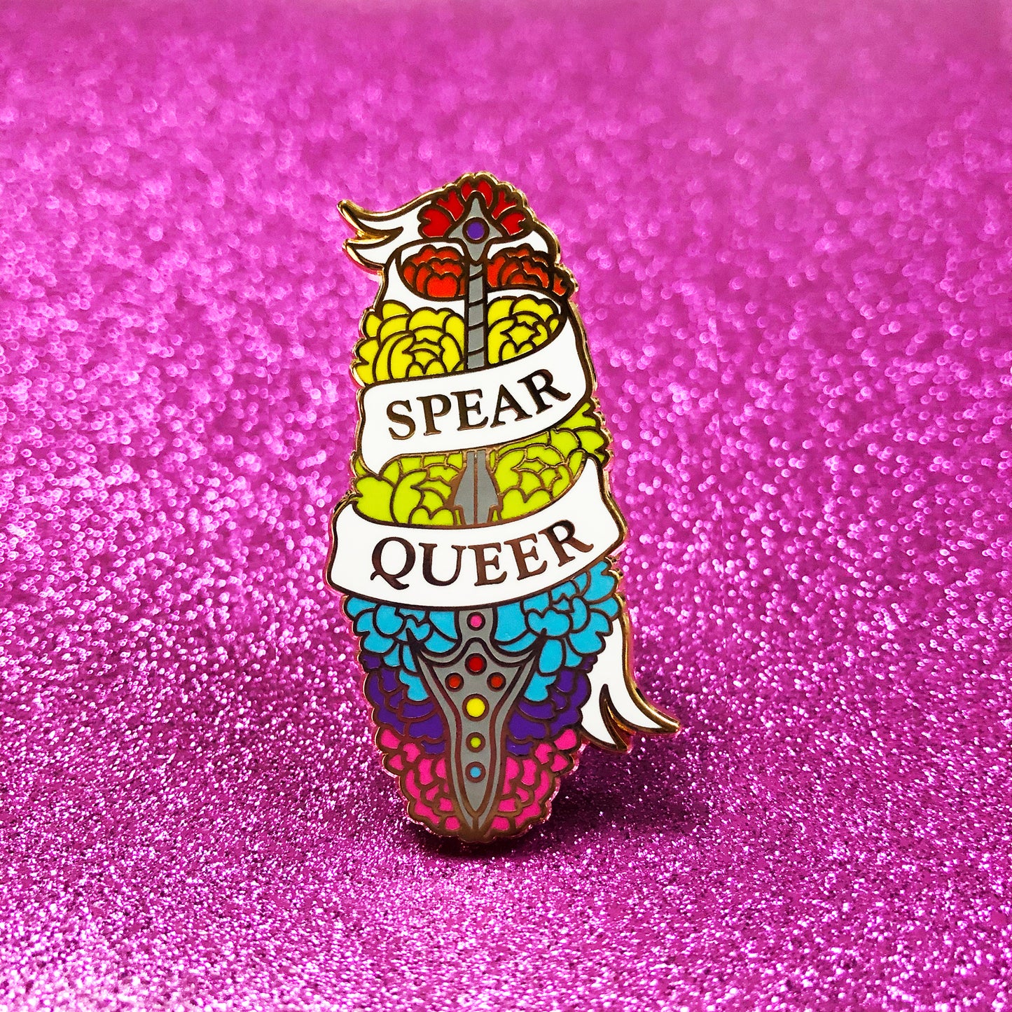 The goldcast pin features a decorative spear in a bed of flowers in the rainbow hues of the pride flag. A ribbon trails across the front and reads "Spear Queer."