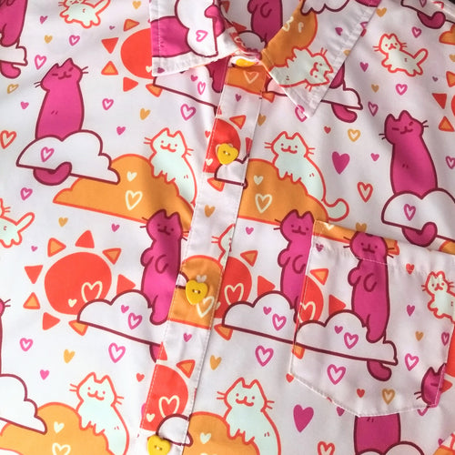 A soft pink button up shirt with yellow heart shaped buttons. The shirt pattern features cute white and magenta cats, orange clouds, orange suns, and many purple, orange, and yellow hearts. 