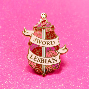 The goldcast pin features an ornate sword in a bed of roses in the orange and pink colors of the lesbian flag. A ribbon trails across the front and reads "Sword Lesbian."