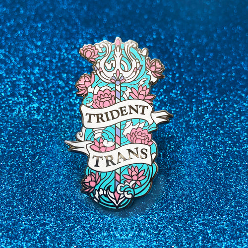 The goldcast pin features an ornate trident in a bed of flowers and rushing water in the pastel blue, white, and pink colors of the trans flag. A ribbon trails across the front and reads 