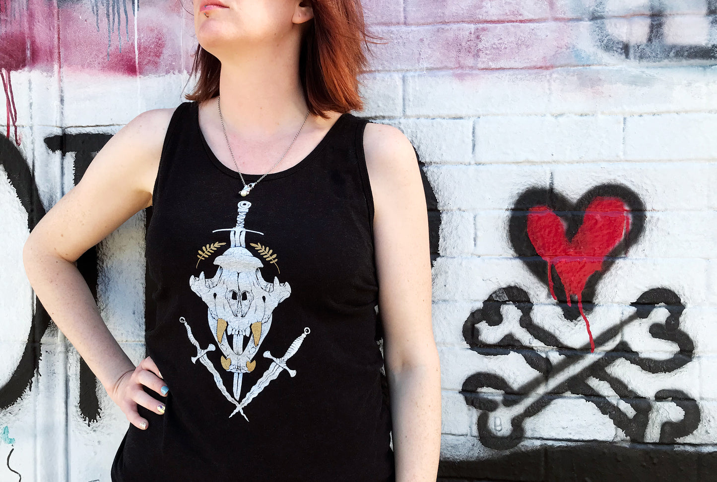 Black tank top with white tiger skull and daggers on model in front of graffiti wall.