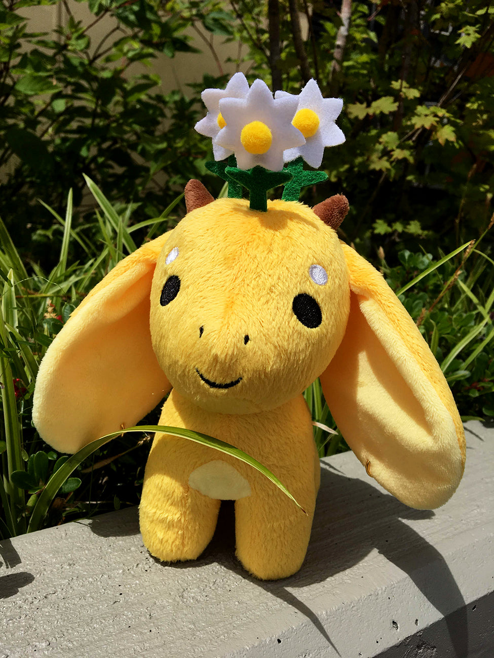 Chamomile plush stands outside in the sun in front of greenery.
