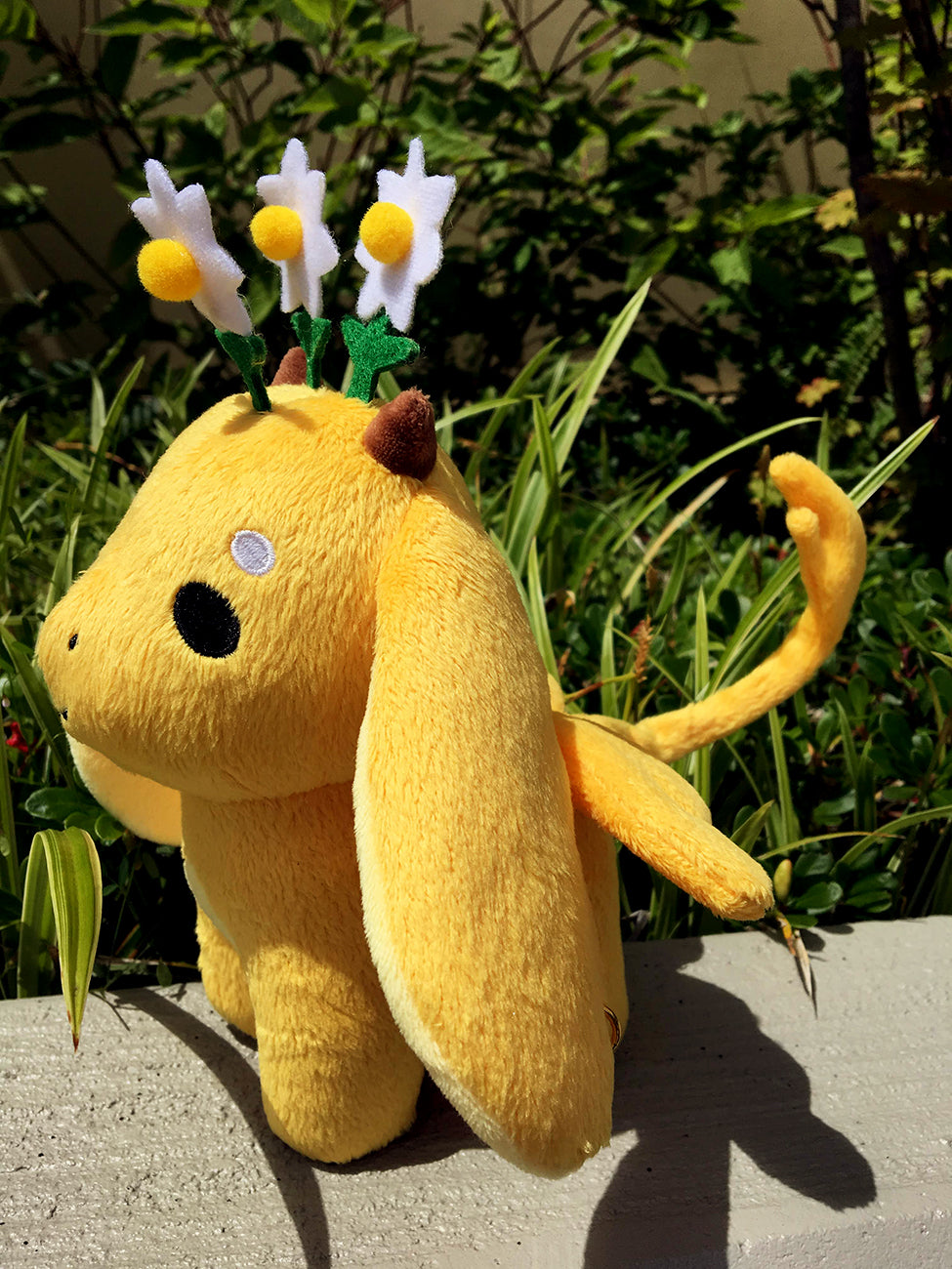 Side view of chamomile plush standing in the sun in front of greenery.