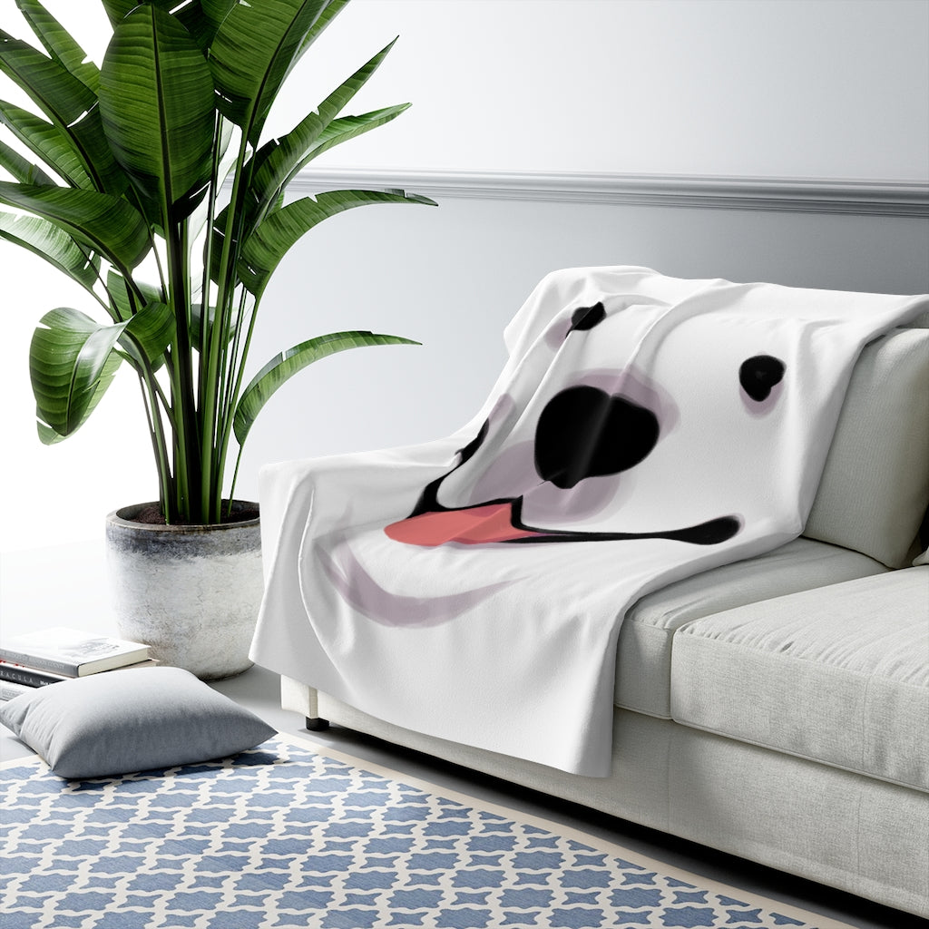 White blanket with closeup of Cloud the dog's face with pink tongue out draped over light grey couch next to palm plant.