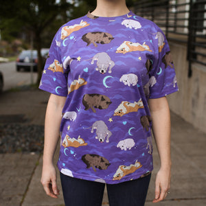 Model posing on sidewalk wearing dark blue jeans and purple t-shirt with cloud, crescent moon, heart, and star motifs surrounding 4 different sleeping dog breeds..