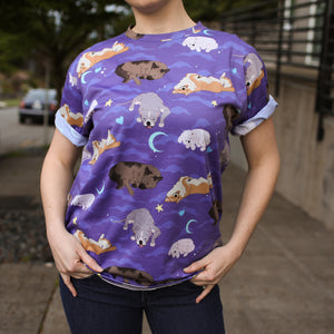 Model posing on sidewalk wearing dark blue jeans and purple t-shirt with cloud, crescent moon, heart, and star motifs surrounding 4 different sleeping dog breeds..