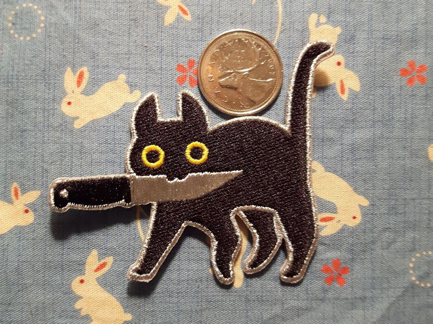 Embroidered patch depicting a black cat with yellow eyes holding knife in mouth on light blue and white rabbit patterned cloth. Canadian quarter for scale..