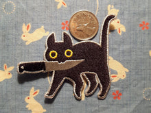 Load image into Gallery viewer, Embroidered patch depicting a black cat with yellow eyes holding knife in mouth on light blue and white rabbit patterned cloth. Canadian quarter for scale..