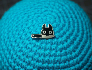 Silver enamel pin of black cat head holding a knife in its mouth on knitted light blue pouf.