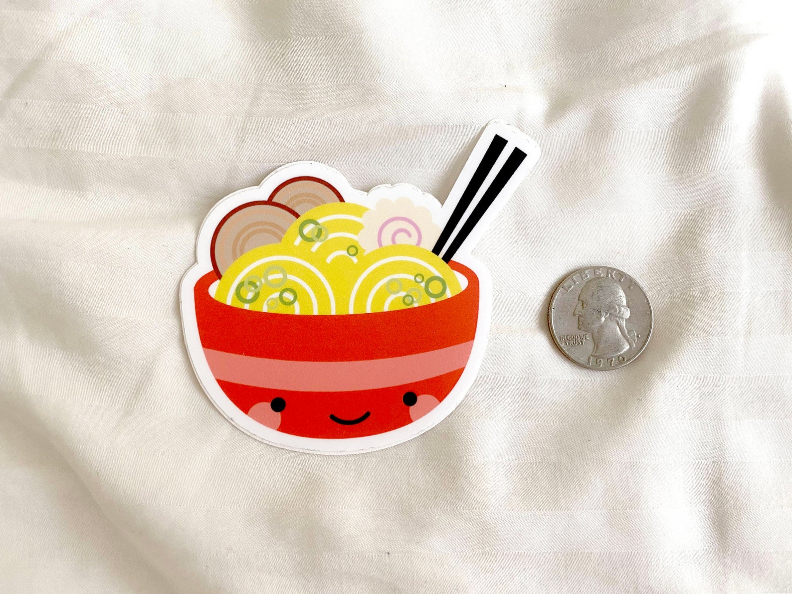 One ramen sticker next to quarter for scale on a white cloth background.