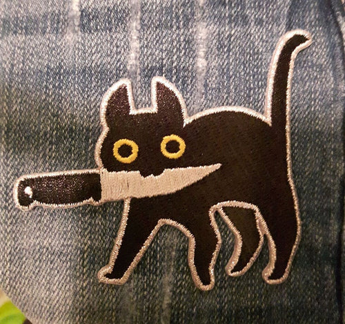 Embroidered patch depicting a black cat with yellow eyes holding knife in mouth on light wash denim.