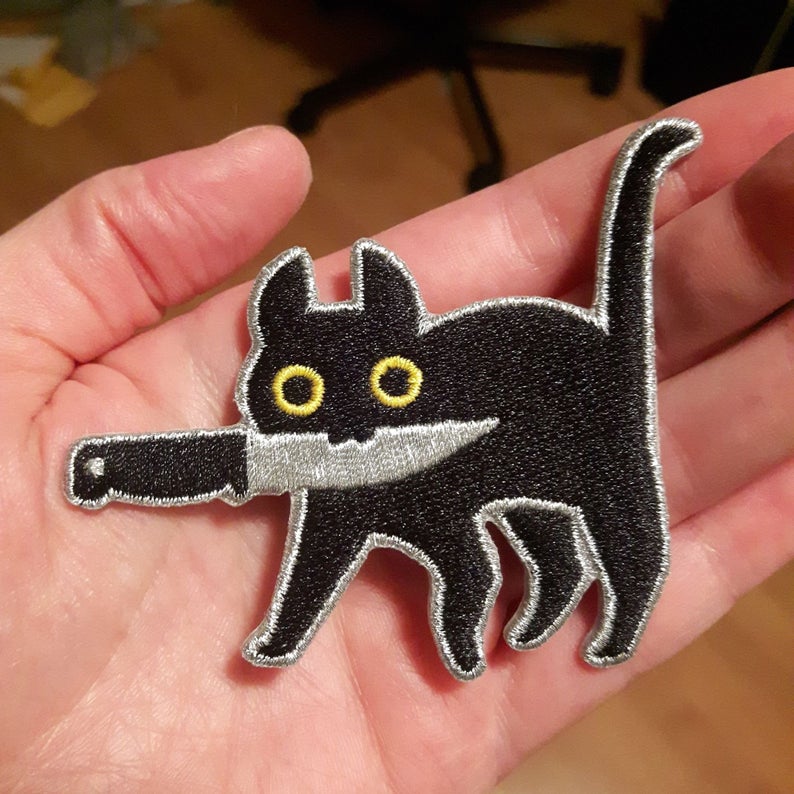 Embroidered patch depicting a black cat with yellow eyes holding knife in mouth in model's hand.