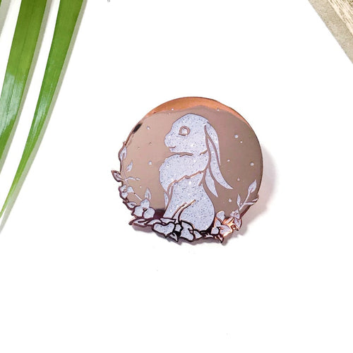 Sparkly white and gold enamel pin depicting sitting rabbit with flowers. 