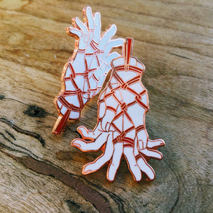 Two rose gold pins depicting hands tenderly tied with rope on wooden table.