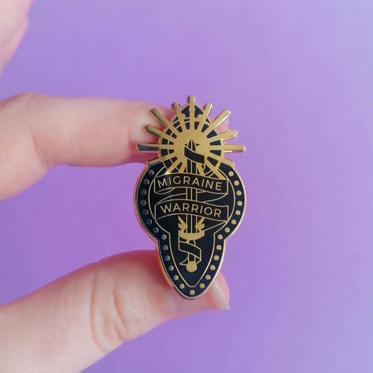 Model holds a black and gold enamel pin with design depicting a ribbon wrapped around a sword and sun, reading "Migraine Warrior."