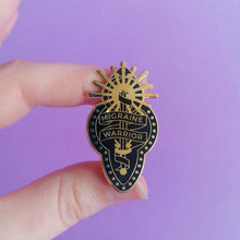 Load image into Gallery viewer, Model holds a black and gold enamel pin with design depicting a ribbon wrapped around a sword and sun, reading &quot;Migraine Warrior.&quot;